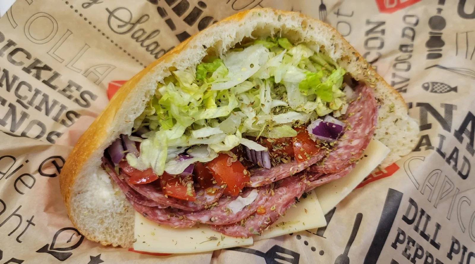 A submarine sandwich with lettuce, tomatoes, onions, slices of cheese, and salami on a piece of paper.
